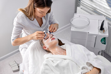 Obraz na płótnie Canvas attractive woman looking at camera while laying on couch being treated by cosmetologist removing cosmetology mask on face, side view portrait. Professional confident caucasian beautician at work