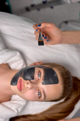 Top View on Young beautiful woman in spa salon with black mask on face, cropped unrecognizable master beautician applied mask on face of young female to cleanse the pores of skin. Facial skin care