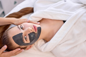 Young caucasian woman uses the services of professional beautician at at spa. cropped unrecognizable master beautician puts black mask on client's face and gives massage. Beauty and health