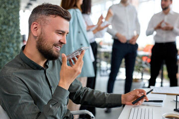 cheerful business worker guy holding mobile phone in hands, talking and listening audio message. excited male in green formal shirt smiling, at break time, have rest. modern technologies, business