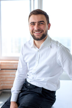 Excited Businessman Or Manager Sit On Desk Posing In Office At Work Place Having Rest, Taking A Break After Business Meeting Or Conference, Looking At Camera Laughing, Wearing Formal Suit