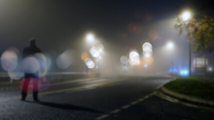 Fototapeta na wymiar A mysterious figure standing in the middle of the road on a misty night. With a blurred, bokeh, out of focus edit