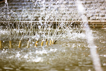Splashing water in the fountain at summer city public urban park. Water jets abstract image close up. Thin jets of pure clear aqua splashes flying upwards under pressure. Gray stone wall background.