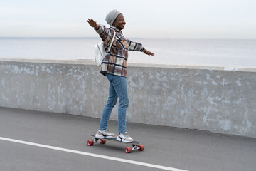 Freedom and urban lifestyle concept: young casual girl skateboarding on longboard at city road near sea coast. Happy millennial active african american woman enjoy extreme leisure activity outdoors.