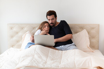 Happy young couple on bed watching scary movie on computer laptop. Young couple in love at home, sitting on bed looking at laptop computer. Young couple lying on cozy bed, looking at computer screen.