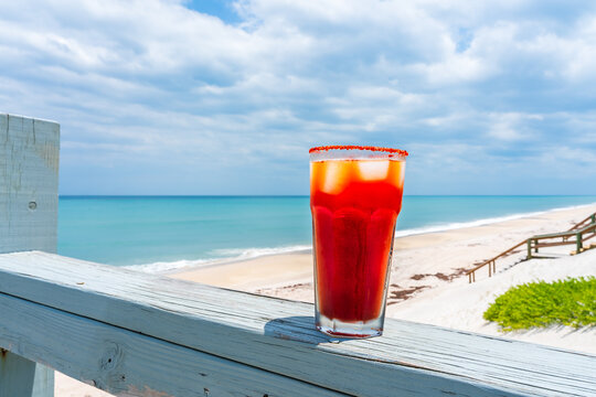 A bright red beer Michelada cocktail in salty-rimmed glasses, glowing in the sun on the veranda railing of a private oceanfront home for brunch