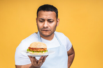 Young african american indian black man eating hamburger isolated over yellow background. Cook preparing burger.