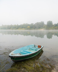 Landscape of Sava river in autumn, moored green and blue boat in shallow water surrounded with algae during beautiful misty morning