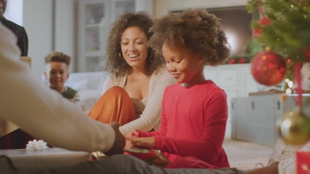 Girl unwrapping present as multi-generation family exchange gifts around christmas tree at home - shot in slow motion