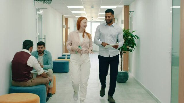 A handsome black man is walking with a stunning ginger woman, they are work colleagues and the black man is holding a tablet talking about work with the woman, they are sitting at a modern office
