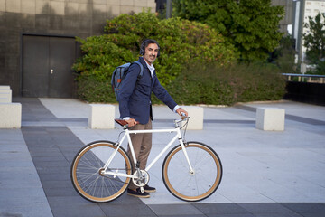 Bearded businessman, handsome and self-confident, carrying a backpack, walking outdoors with a bicycle. Eco-friendly transportation concept