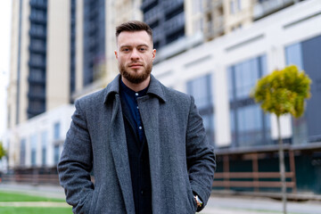 Naklejka premium Outdoor fashion male portrait. Handsome and charming young businessman dressed in suit and coat standing outside a modern building. Head and shoulders portrait