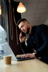 Confident businessman discusses issues on the phone and using laptop in a cafe. Busy man working during coffee break. Closeup