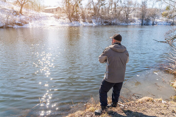 Fototapeta premium A fisherman with a fishing rod catches fish on the banks of a snow-covered river in early spring