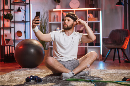 Happy bearded man in sport clothes sitting on floor and taking selfie on smartphone. Middle aged male taking fun and relaxing after domestic workout during evening time.