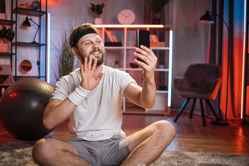 Caucasian bearded man in sport clothes waving and smiling during video call after domestic workout. Middle aged person sitting on floor at living room. Concept of sport and technology.