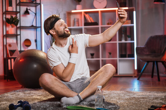 Middle aged bearded man with funny face using modern smartphone for taking selfie. Caucasian male in sportswear, headband and socks resting on floor after training.