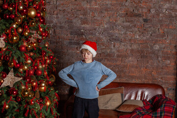 dissatisfied boy in hat stays by Christmas tree . Boy doesn't get presents for christmas. the parcel with gifts did not arrive from online shop. Kid is without presents for Christmas.