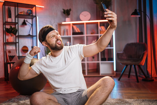 Leisure and recreation at home concept. Joyful handsome smiling young bearded guy in sportswear sitting on the floor with crossed legs, making selfie demonstrating his biceps and showing tongue