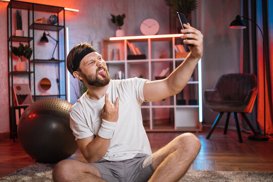 Bearded man showing horns rock gesture and tongue out while taking selfie on smartphone. Bearded male using modern cell phone while relaxing after domestic workout.