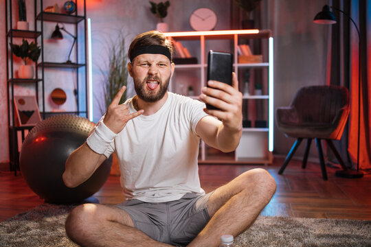 Bearded man showing horns gesture and sticking out tongue while taking selfie on smartphone. Bearded male using modern cell phone while relaxing after domestic workout.