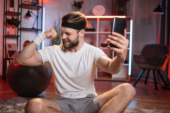 Positive caucasian man in activewear sitting on floor after evening training and taking selfie on modern smartphone, showing his strong arms. Concept of people, sport and technology.