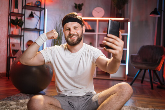 Positive caucasian man in activewear sitting on floor after evening training and taking selfie on modern smartphone. Concept of people, sport and technology.