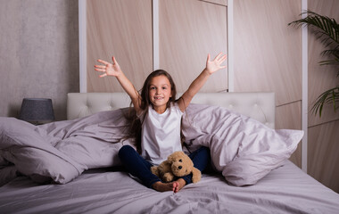 a little brunette girl in pajamas is sitting under a blanket in bed with a teddy bear