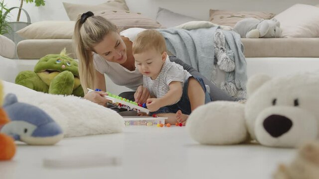 Happy smiling mom plays with her beautiful baby, with colorful educational learning toy, sitting at home between pillows and animals plush, healthy and cared for growth concept