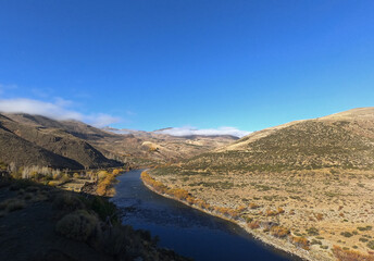 Idyllic rural landscape with a clear blue sky. The river flowing across the golden valley, yellow grassland, forest and mountains in autumn.