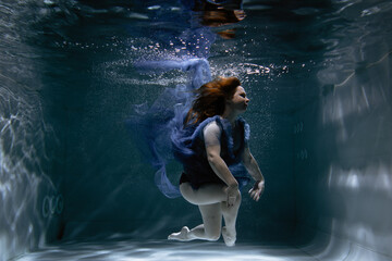 A beautiful girl in a blue dress swims underwater in the pool like a little mermaid from a fairy tale