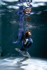 Fototapeta premium A beautiful girl in a blue dress swims underwater in the pool like a little mermaid from a fairy tale