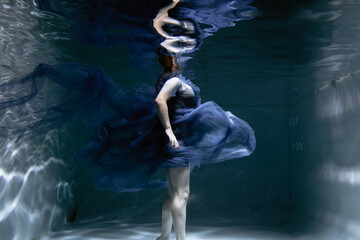 A beautiful girl in a blue dress swims underwater in the pool like a little mermaid from a fairy...