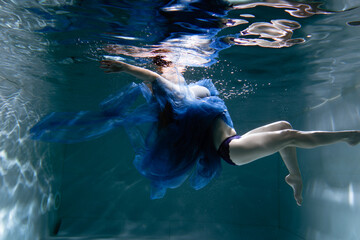 Naklejka premium A beautiful girl in a blue dress swims underwater in the pool like a little mermaid from a fairy tale