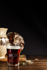 Kvass, a refreshing natural fermented drink. A glass of a dark drink with a dense foam. A ceramic jug and rye bread in the background. Vertical, on a black background