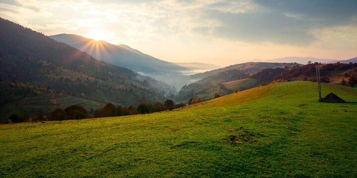 rural landscape at sunrise. beautiful autumnal mountain scenery. green grassy meadow on the hillside. fog down in the valley. sun and fluffy clouds above horizon