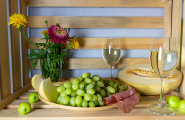 Green grape, ripe melon, meat, white wine, apples, red and yellow flowers in wooden box background