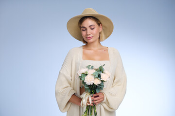 A young beautiful blonde caucasian woman with a short haircut in a beige suit and hat with bouquet of white flowers on blue background. Beauty portrait. The girl smiles, clean skin and white teeth.