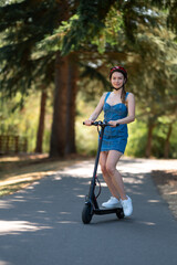 A cute slender girl rides an electric scooter in the park. Summer sunny day. Holidays, outdoor activities, care for the environment, sustainable transport, transport of the future.