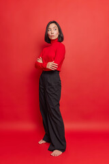 Pensive Asian woman stands in full length keeps arms folded looks thoughtfully wears turtleneck...