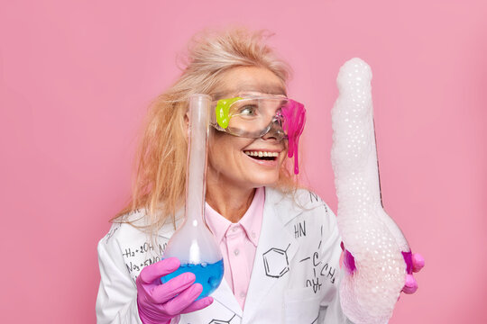 Happy research scientist holds glass flasks makes experiment look at reaction with bubbles after mixing reagents glad after solution distillation result wears safety glasses white coat poses indoor