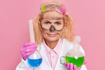 Discontent professional female chemist has shaggy hair dirty face after explostion of substance holds glass flasks with colorful liquid dressed in white coat safety glasses on forehead poses indoor