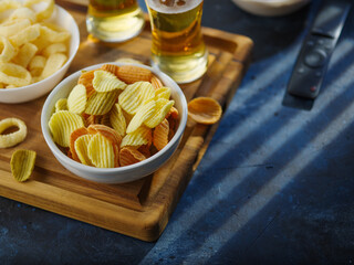 Snacks, chips and light beer in glasses, TV remote control. High angle view. Dark blue background. Watching TV programs, sports programs with your family, with friends.