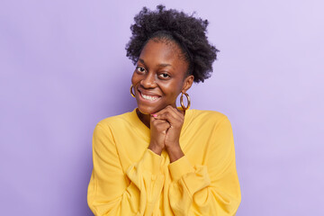 Fototapeta na wymiar Portrait of good looking dark skinned young woman with natural curly hair keeps hands together near face smiles toothily feels touched wears casual yellow jumper isolated over purple background.