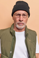 Obraz premium Serious fashionable bearded man looks directly at camera wears round spectacles hat and vest listens attentively information isolated over beige background. Strict dissatisfied elderly male model