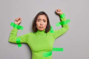 Upset Asian woman with dark hair has gloomy sulking expression plastered with tapes to wall being...