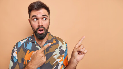 Shocked bearded man shows unexpected revelation indicates at promo shows copy space wears colorful shirt isolated over beige background gasps overwhelmed stares surprised. Omg look at this advert