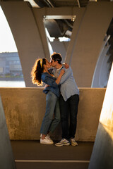 Sensual romantic couple of young adult male and female happy embracing and kissing at sunset standing under bridge in urban space. Tender hug of man and woman in love. Romance and relationship concept