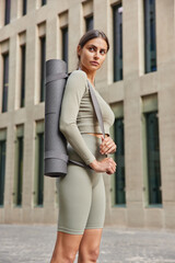 Vertical shot of thoughtful slim woman in sportsclothes carries rolled karemat going to have sport training focused back into distance poses against urban background. Sporty lifestyle concept