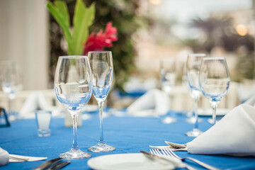 Fototapeta premium Close-up of crystal glasses on a restaurant table with a blue tablecloth, plates, cutlery, napkins and flowers, with a blurred garden in the background at a social event